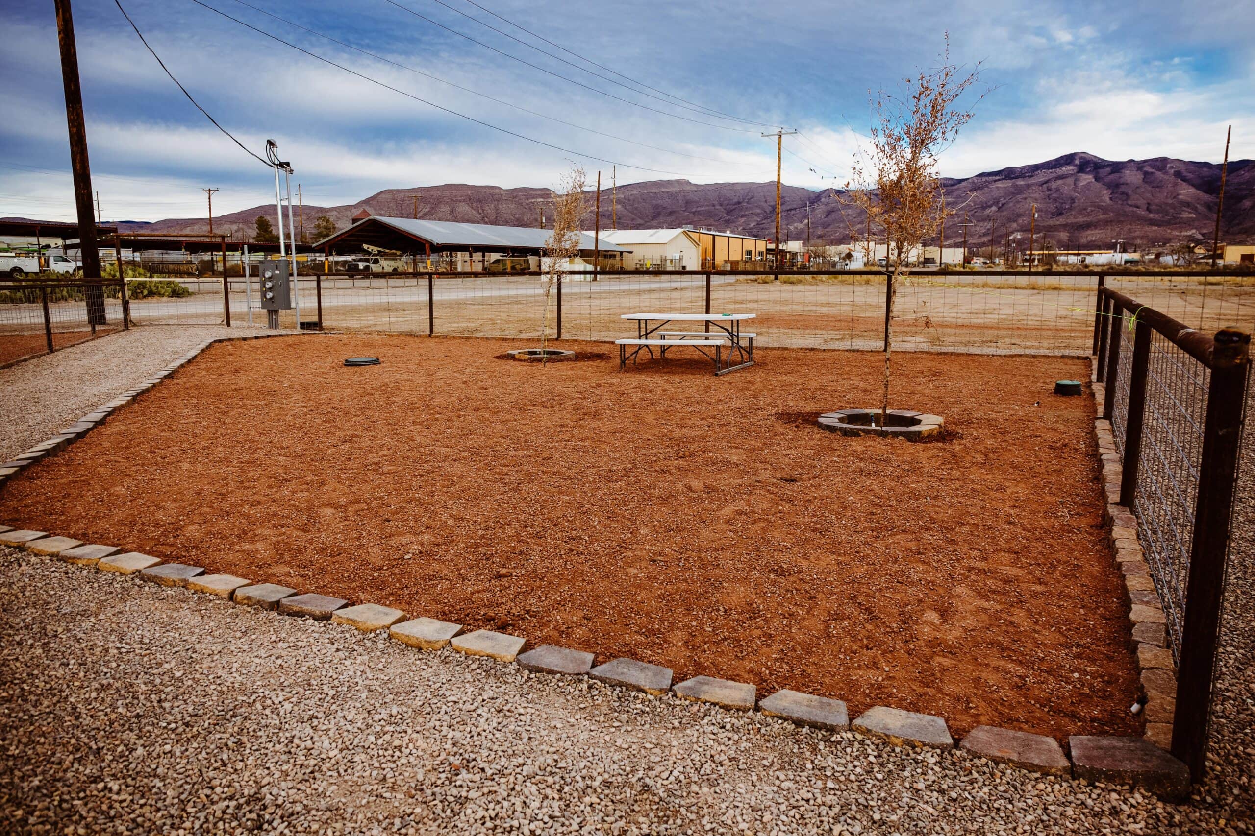 Fenced area with a dirt ground, two small trees, a picnic table, and mountains in the background.