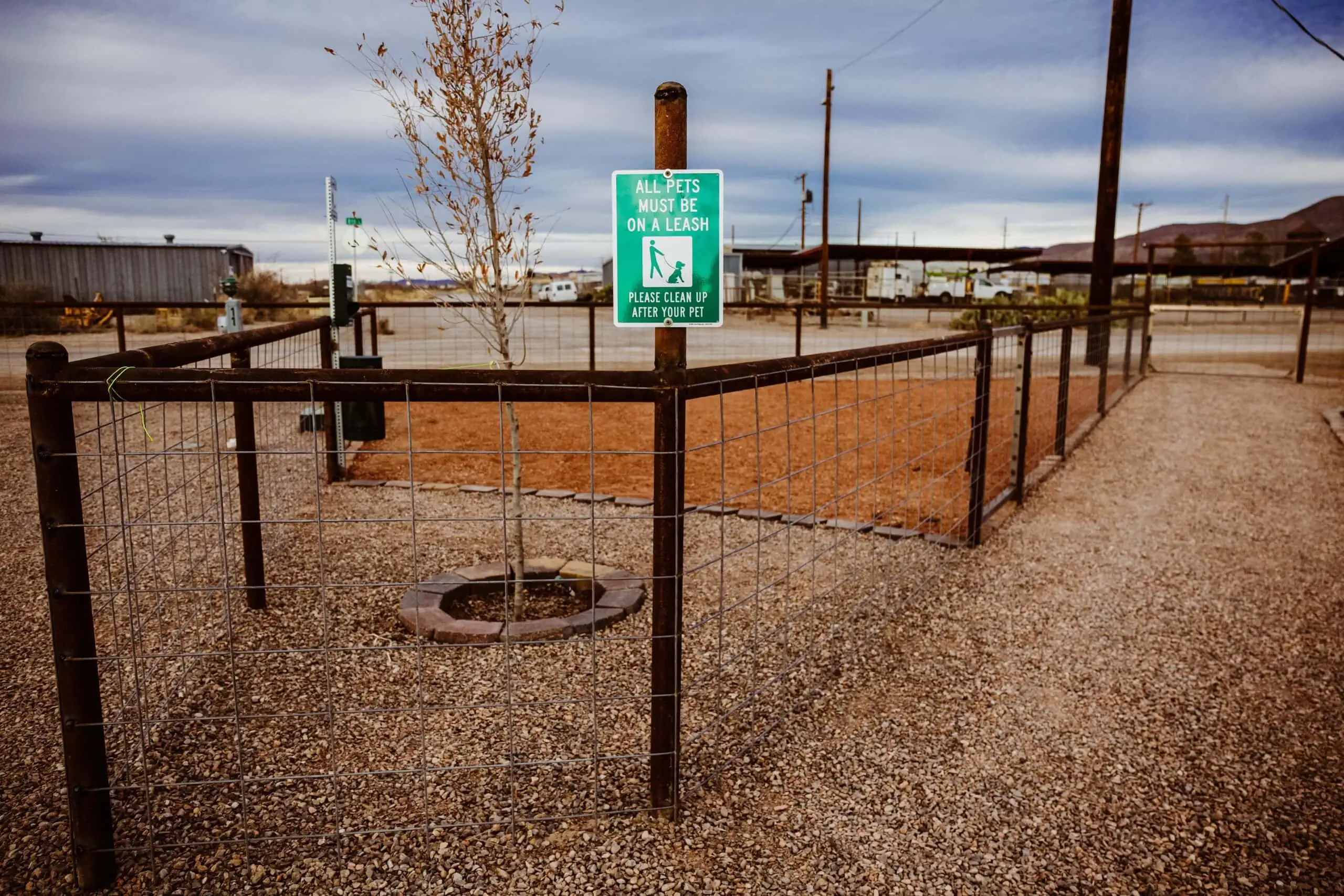 Fenced area with a small tree and a sign reading "All pets must be on a leash. Please clean up after your pet." Surrounding area is gravel, with sparse desert vegetation and buildings in the background.