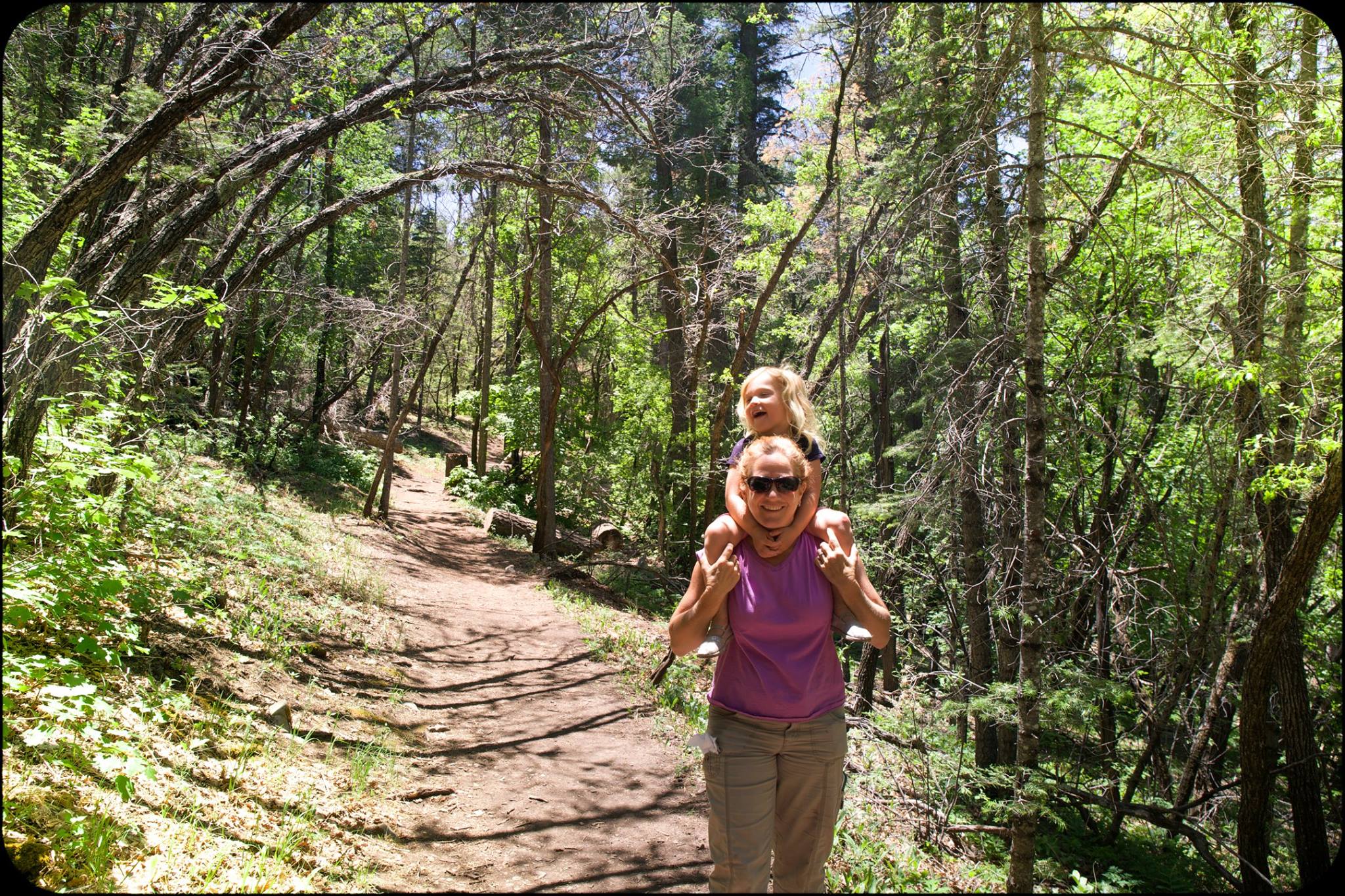 A woman carries a child on her shoulders while walking on a forest path surrounded by trees.