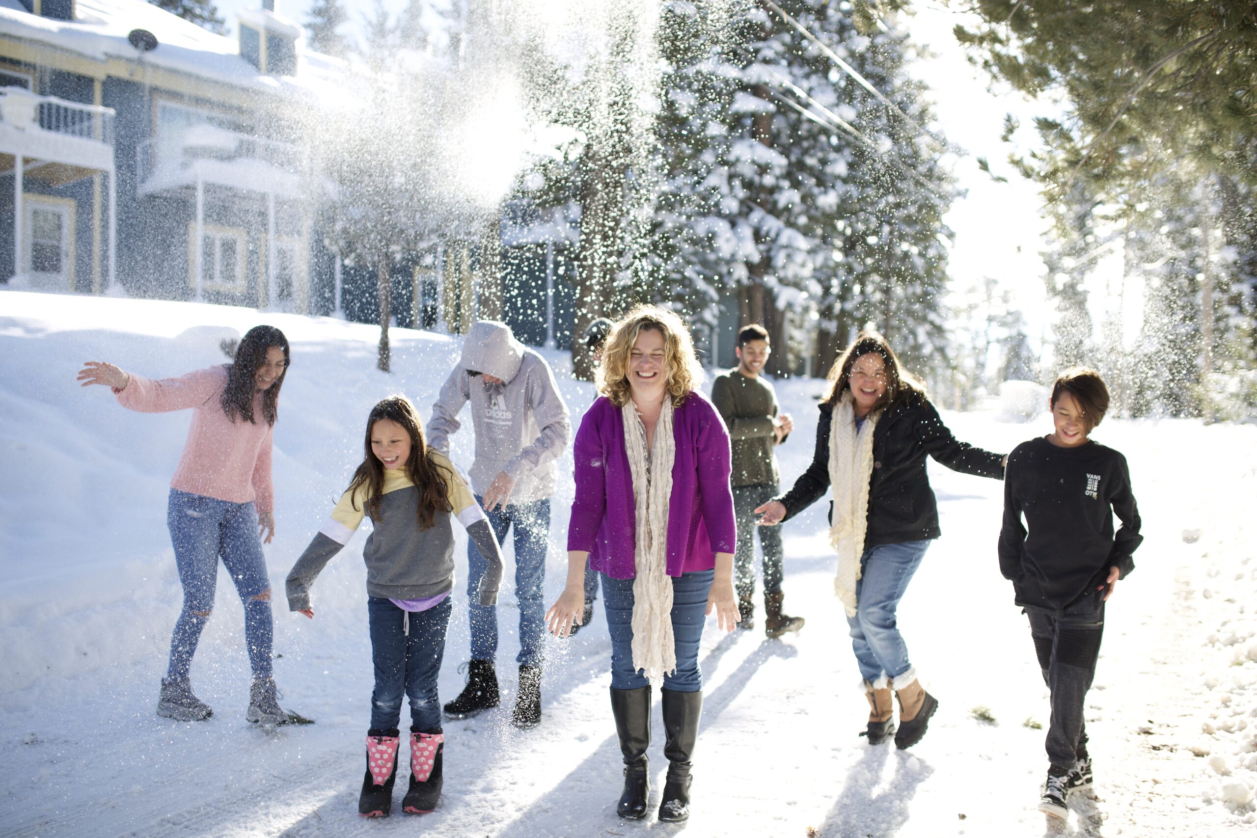 A group of people, including children, play in the snow on a sunny day. They are laughing and throwing snow in the air, with houses and trees covered in snow in the background.
