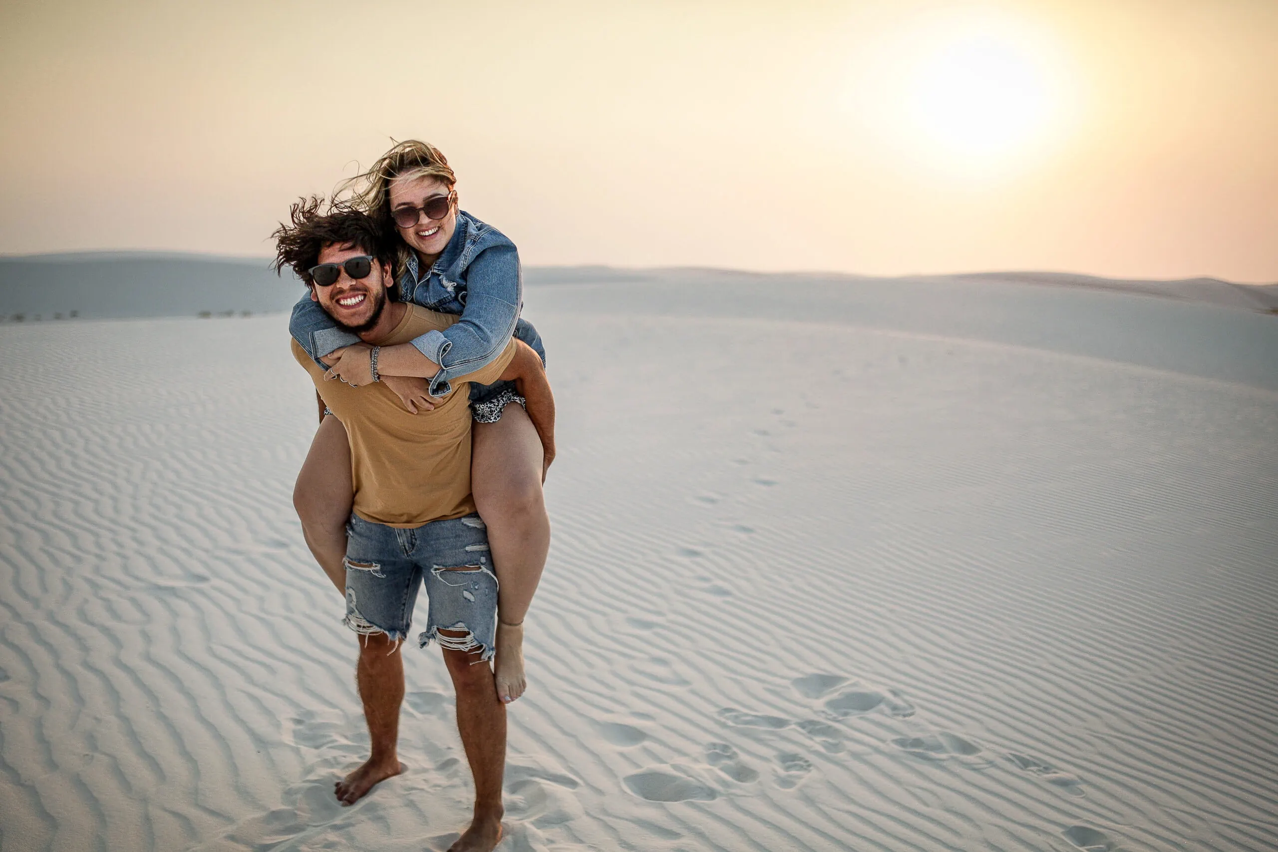 A person carries another on their back while smiling in a sandy desert landscape under a bright sun.