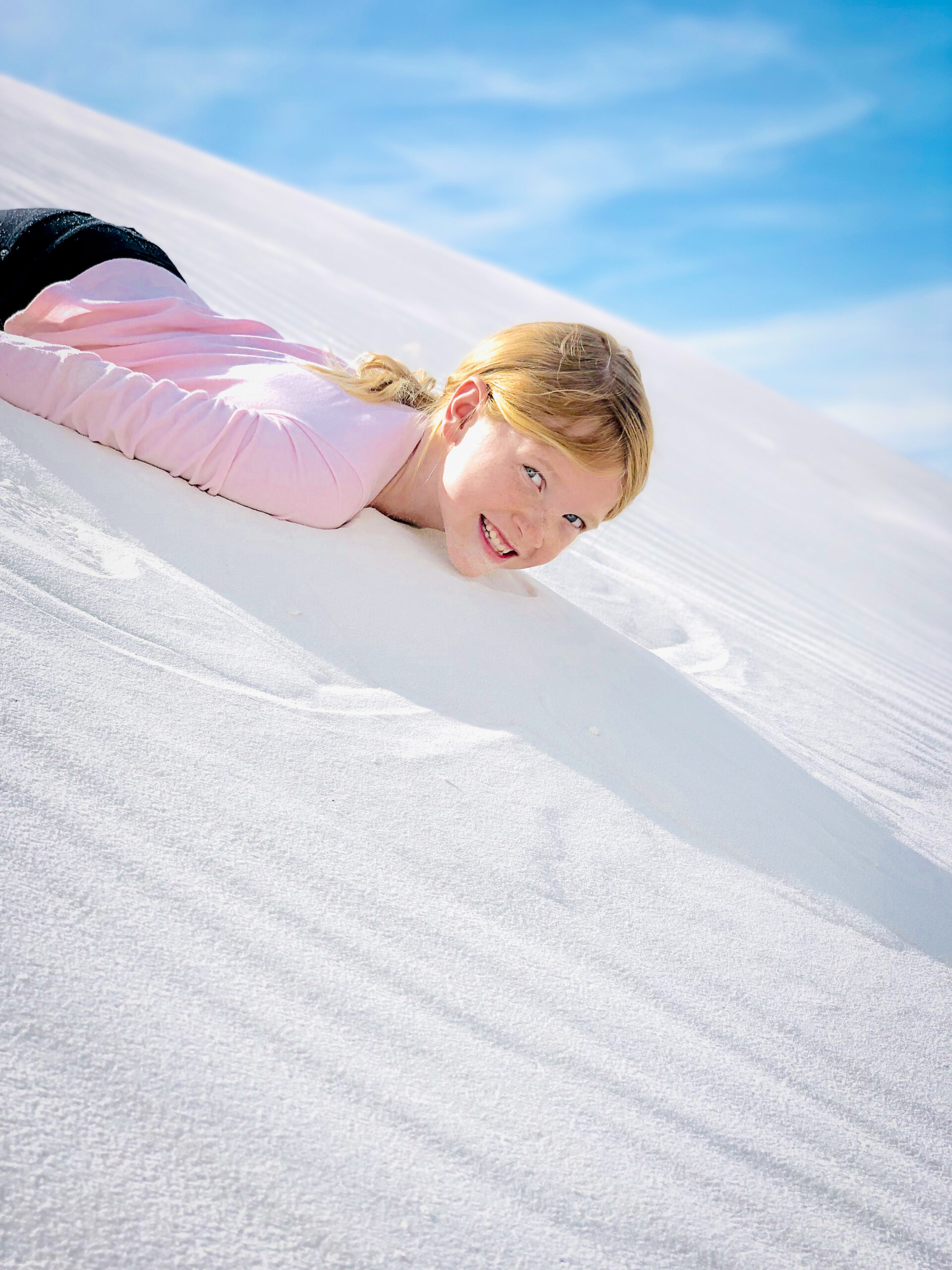 Person lying face-down on white sand under a clear blue sky, smiling at the camera.