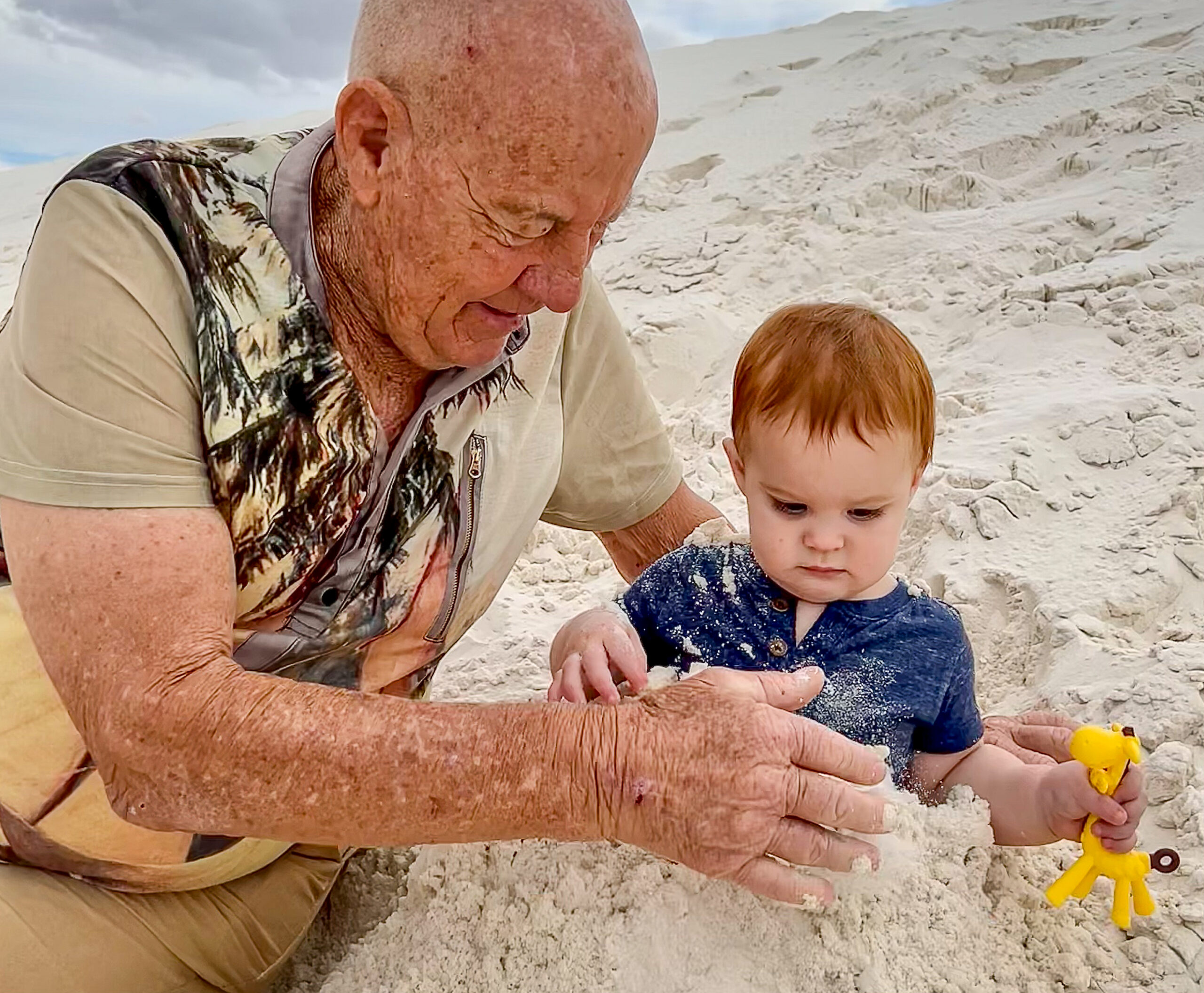 An elderly person and a young child play together in white sand, with the child holding a small yellow giraffe toy.
