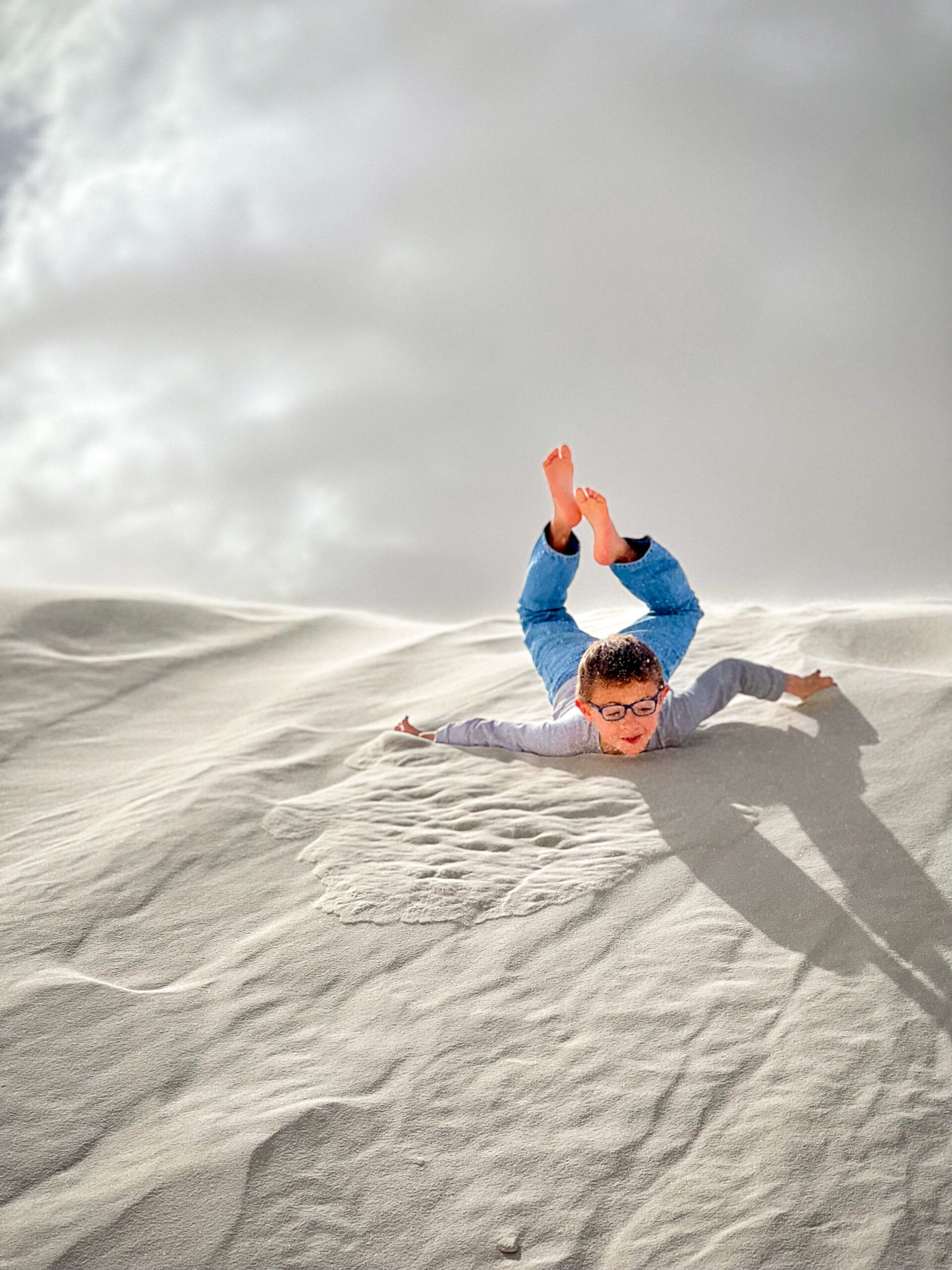 A person slides down a sandy dune under a cloudy sky, feet in the air and arms outstretched.