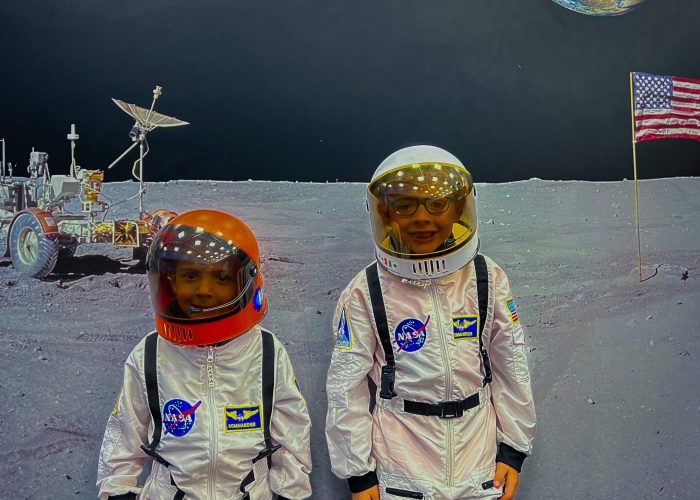 Two children wearing space suits stand in front of a lunar backdrop with the Earth, an American flag, and a lunar rover visible.
