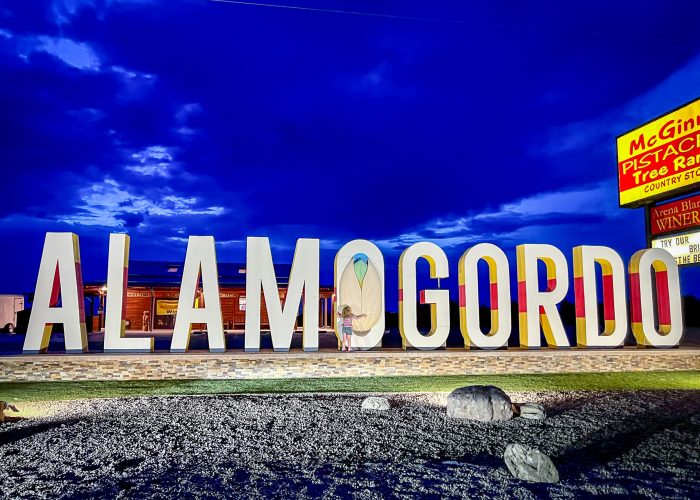 Large illuminated letters spell "ALAMOGORDO" against a dusky sky, with a pistachio tree ranch sign in the background.