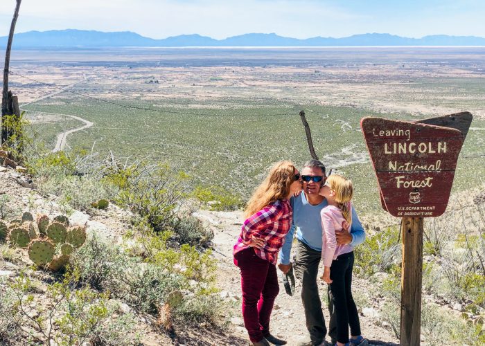A woman and child kiss a man on the cheek next to a "Leaving Lincoln National Forest" sign, with a vast desert landscape in the background.